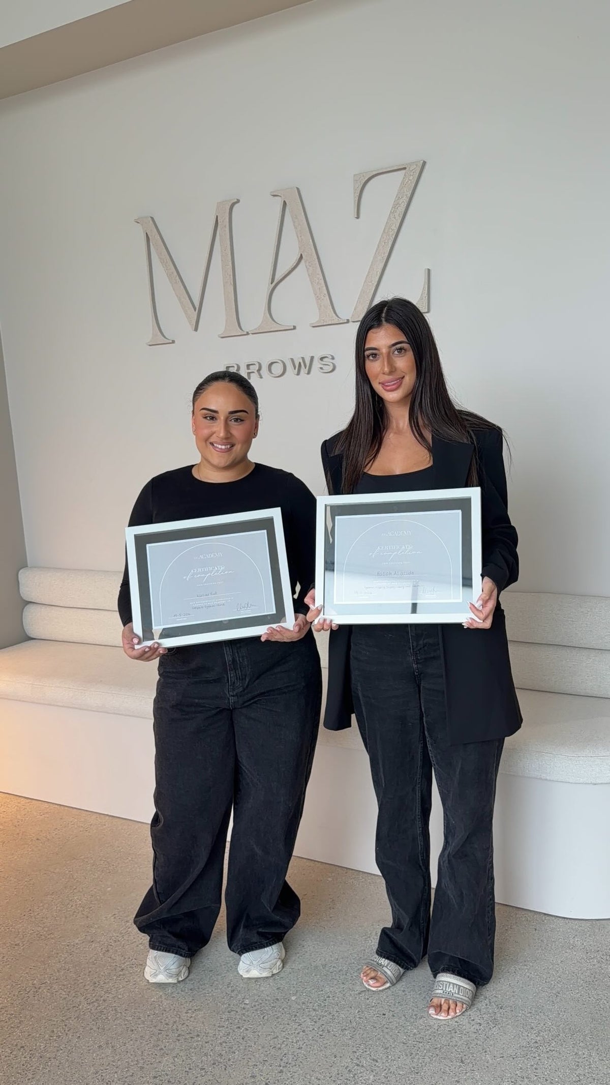 Two women standing indoors in front of a wall with large 'MAZ BROWS' lettering, each holding a framed certificate. Both are dressed in black, and the setting appears to be a salon or beauty studio.